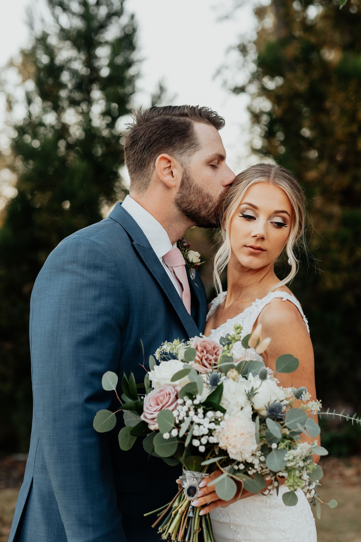 Groom in navy suit kisses bride's temple as she holds a lush bouquet of pink and white flowers with eucalyptus.