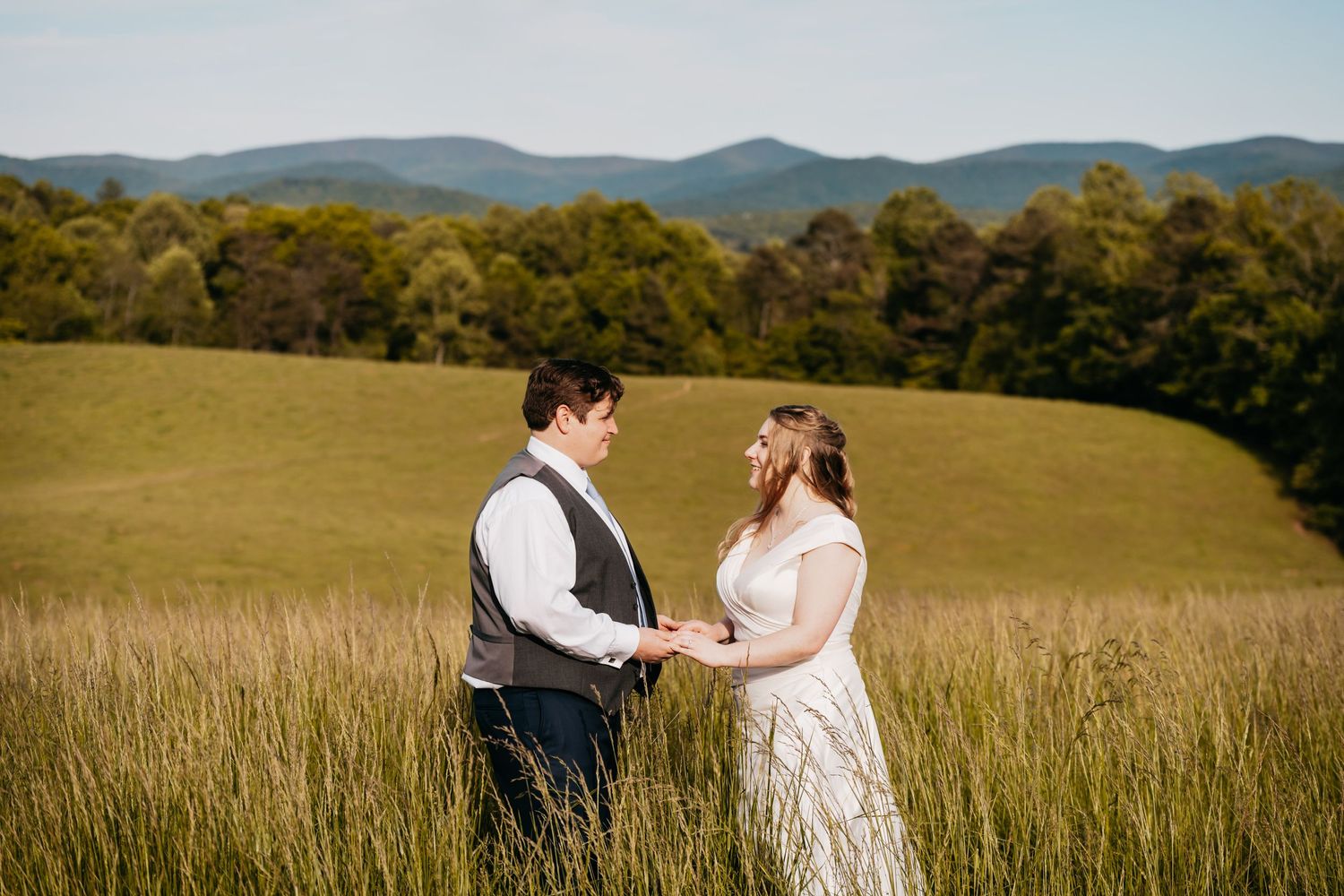 Bride and groom hold hands in a lush green field with rolling mountains in the background.