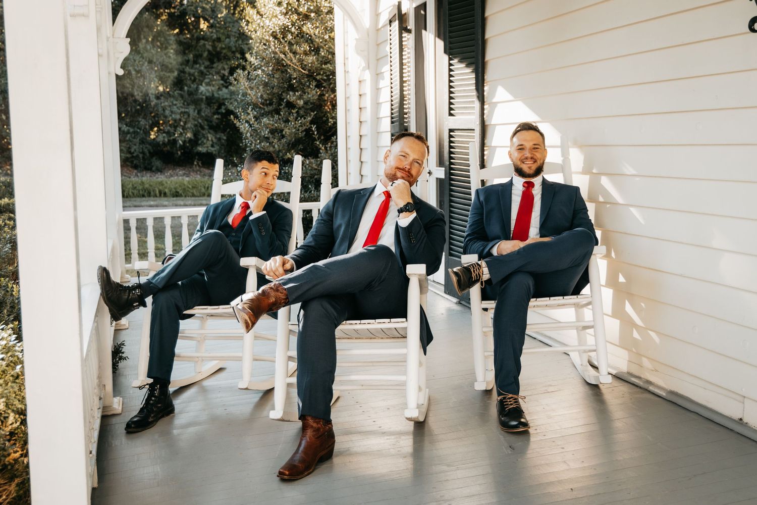 Three groomsmen in navy suits relax on rocking chairs on a classic white porch.