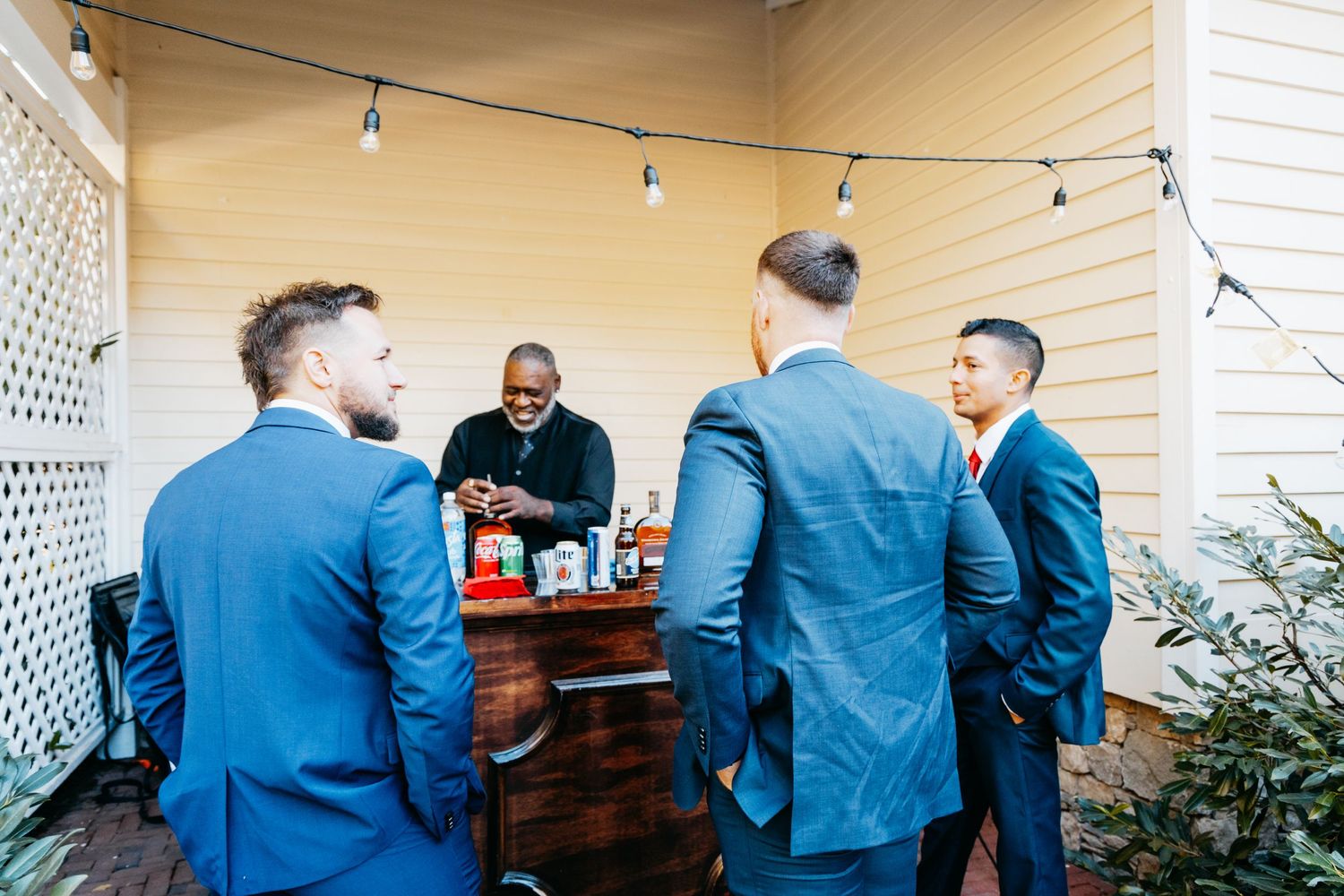 Groomsmen in navy suits gather around a bar cart on a porch before the wedding ceremony.