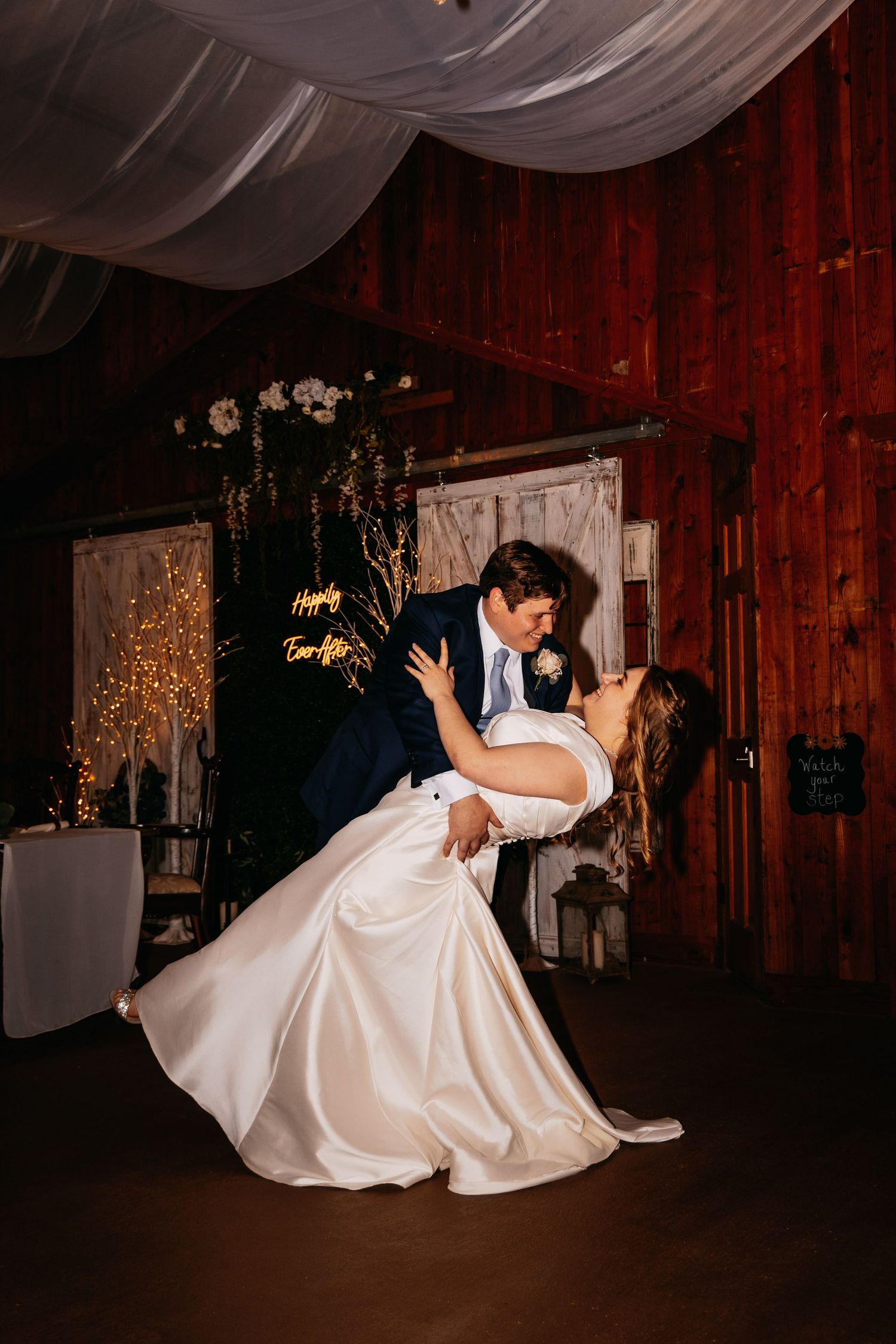 Groom dips bride during first dance in a rustic barn wedding reception with warm string lights.