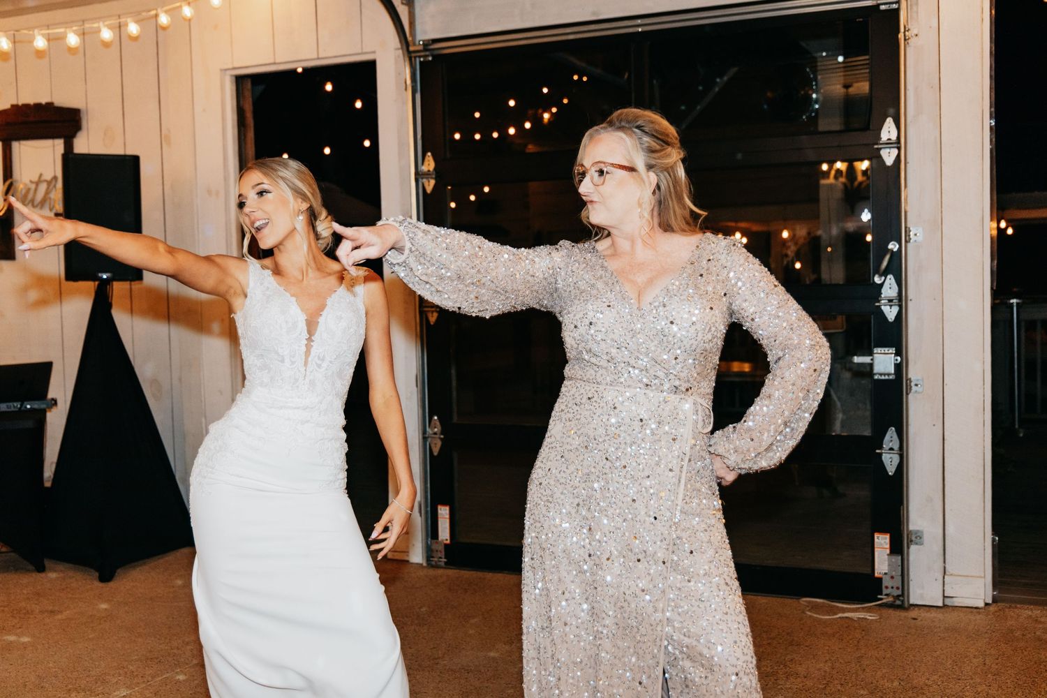 Bride in sleek white gown dances joyfully with a guest in a sequined dress at the wedding reception.