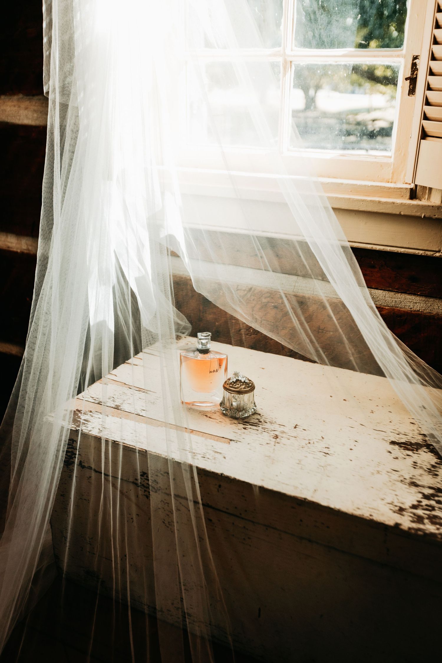 Bridal veil draped over a perfume bottle on a rustic windowsill in soft natural light.
