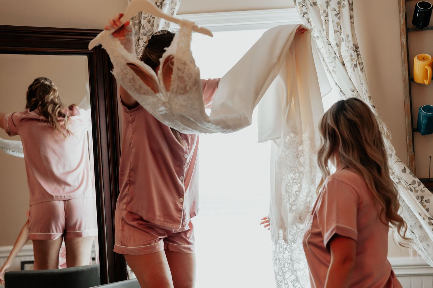 Bridesmaids in pink pajamas helping lift a wedding dress near a bright window.
