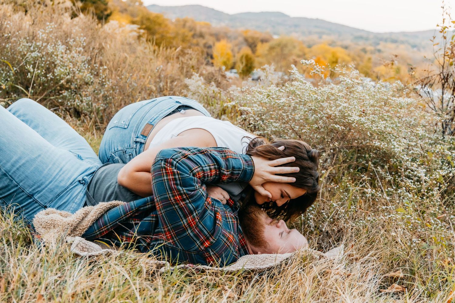 Couple embraces and kisses while lying in a wildflower meadow during autumn engagement session.