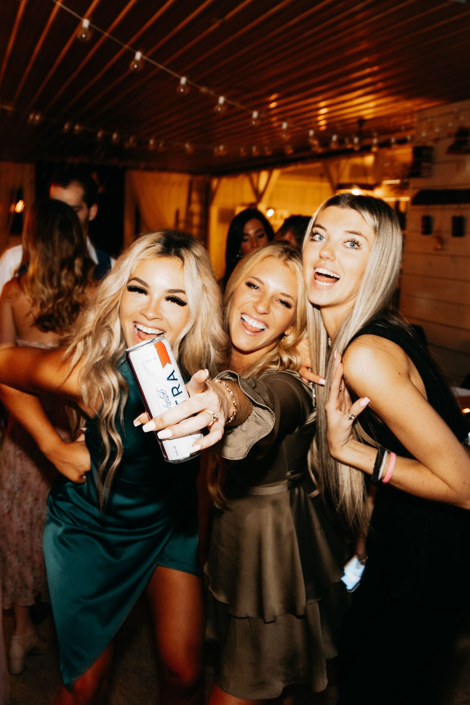 Three women in elegant dresses smile and pose with a drink can during a lively wedding reception party.