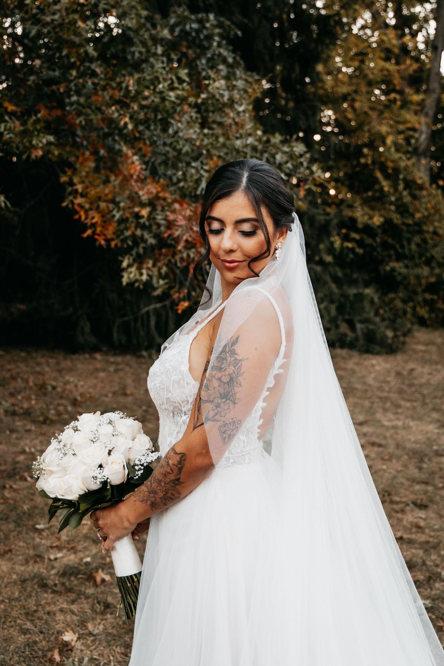 Bride in a lace wedding gown with a long veil holds a white bouquet outdoors.