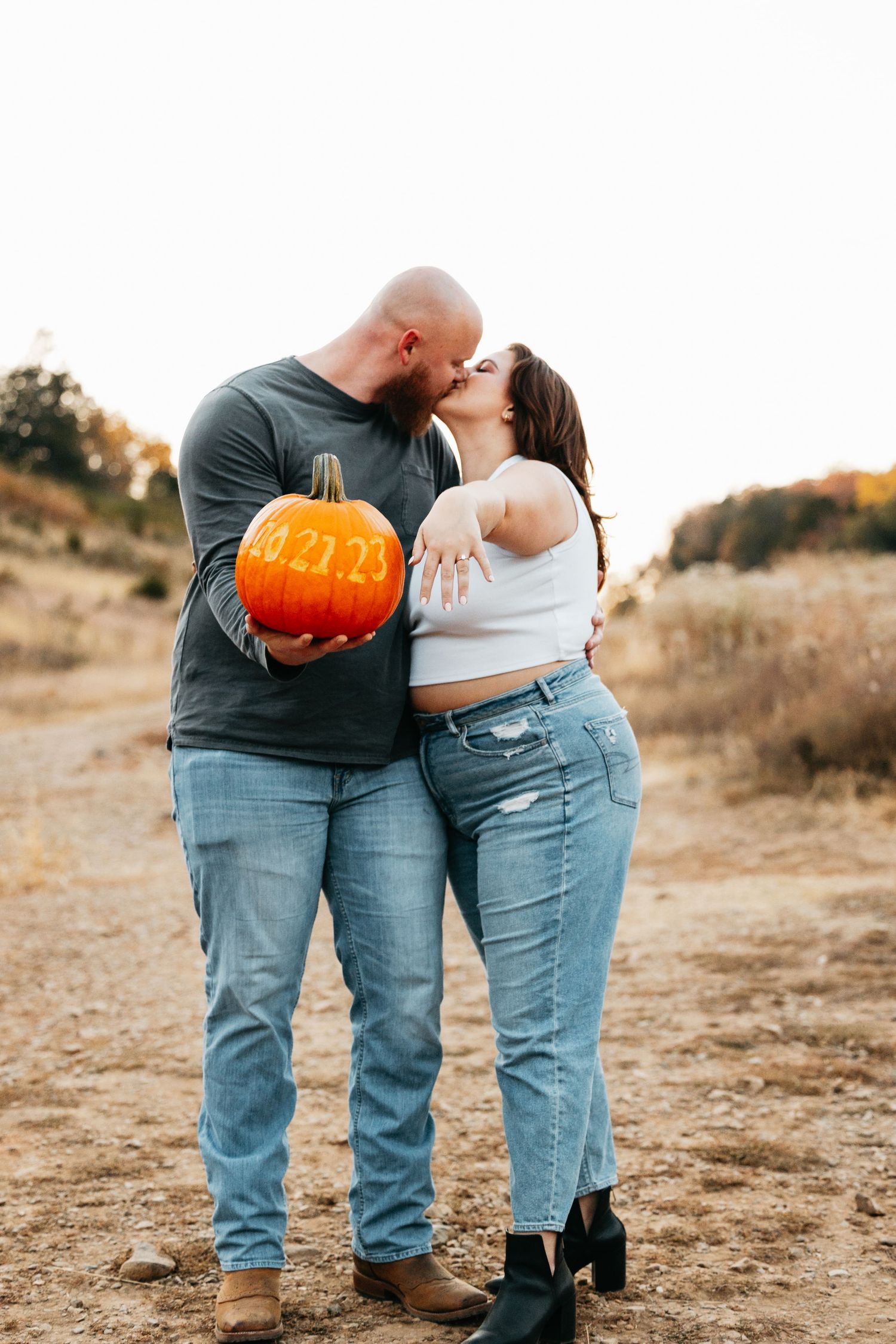 Couple kisses while holding a carved pumpkin with their wedding date during fall engagement shoot.