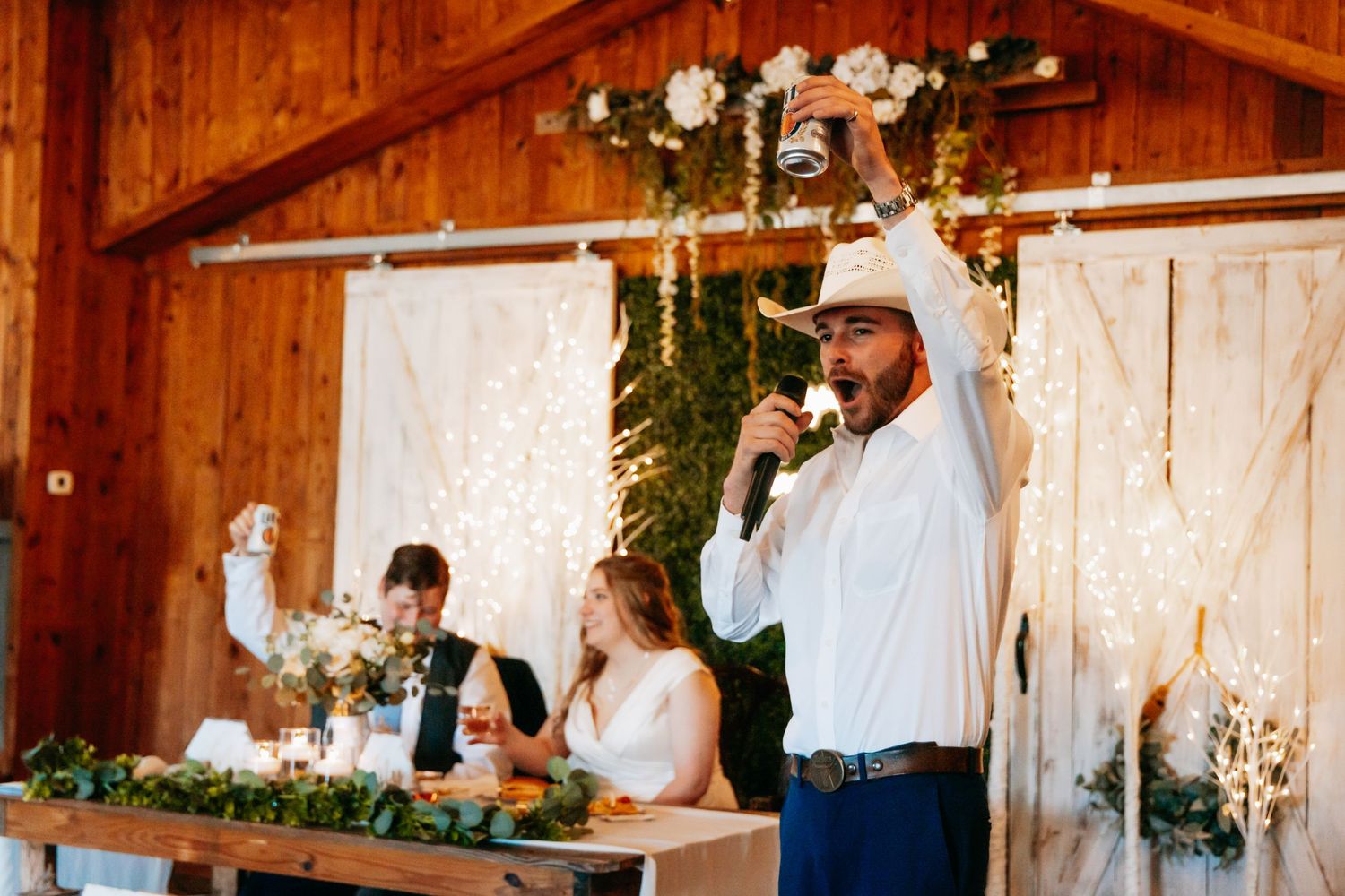 Groomsman in cowboy hat gives a toast at a rustic barn wedding reception with floral decorations.