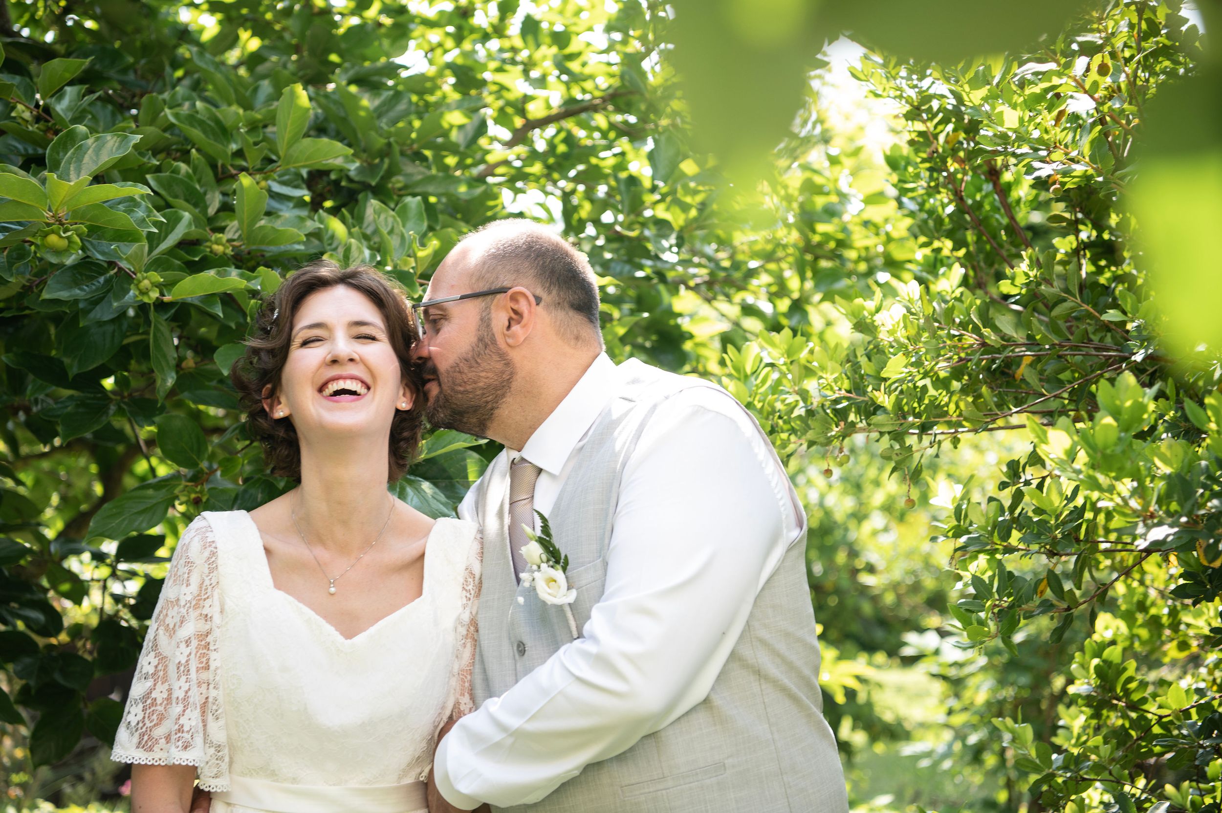 Groom whispers in bride's ear as she laughs. Countryside setting with sunlight in leaves