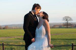Two brides, one in a suit and one in a dress, sit on a fence and kiss in evening light in Wiltshire countryside