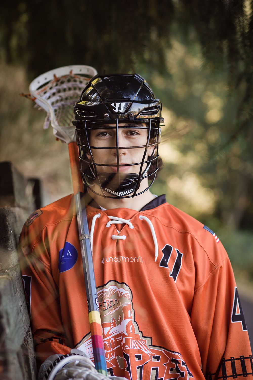 A lacrosse player wearing orange jersey and protective helmet with cage mask stands against blurred outdoor background.