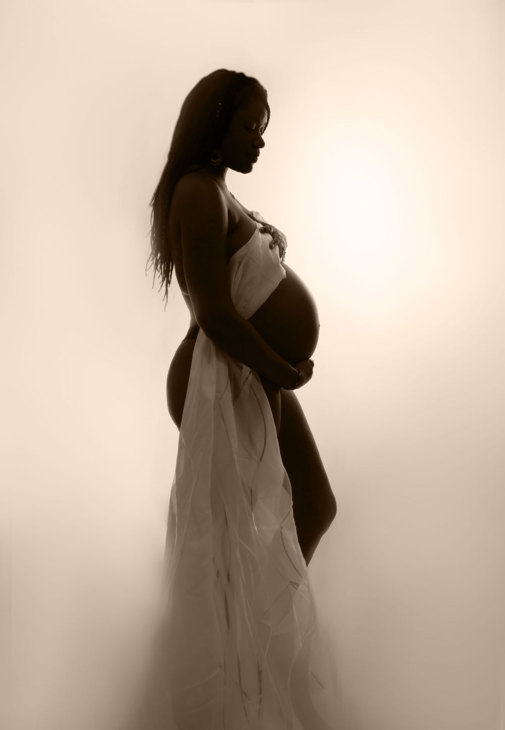Silhouette of an expectant mother in a flowing white dress captured in a dreamy sepia tone.