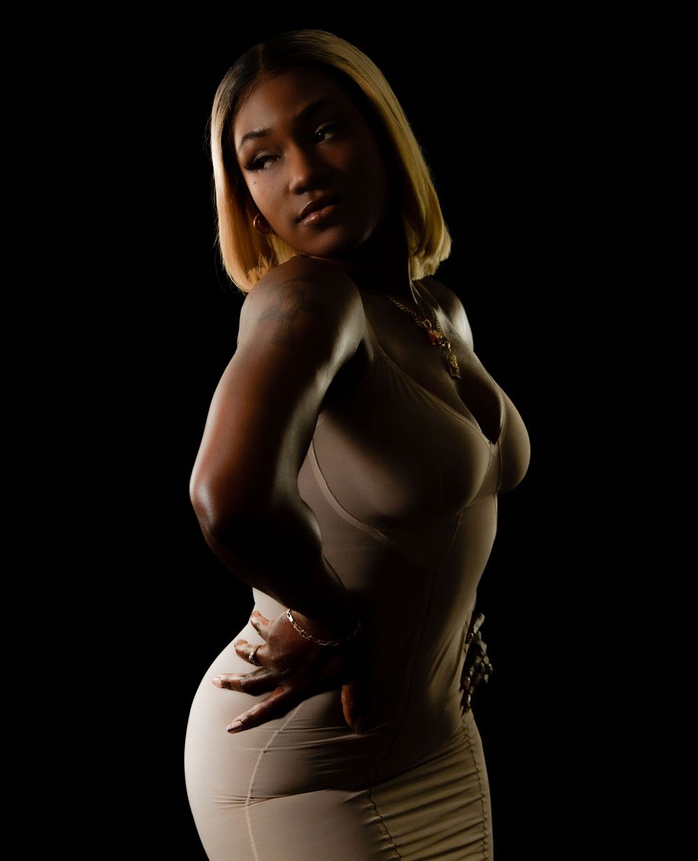 Dramatic low-light portrait in beige fitted dress against dark background.