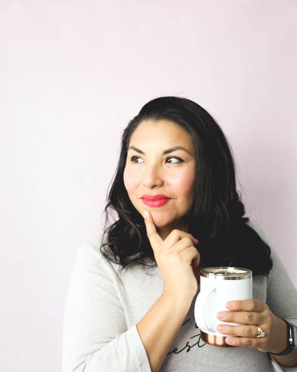 A thoughtful person in a light grey top holds a white mug while posing against a neutral background.