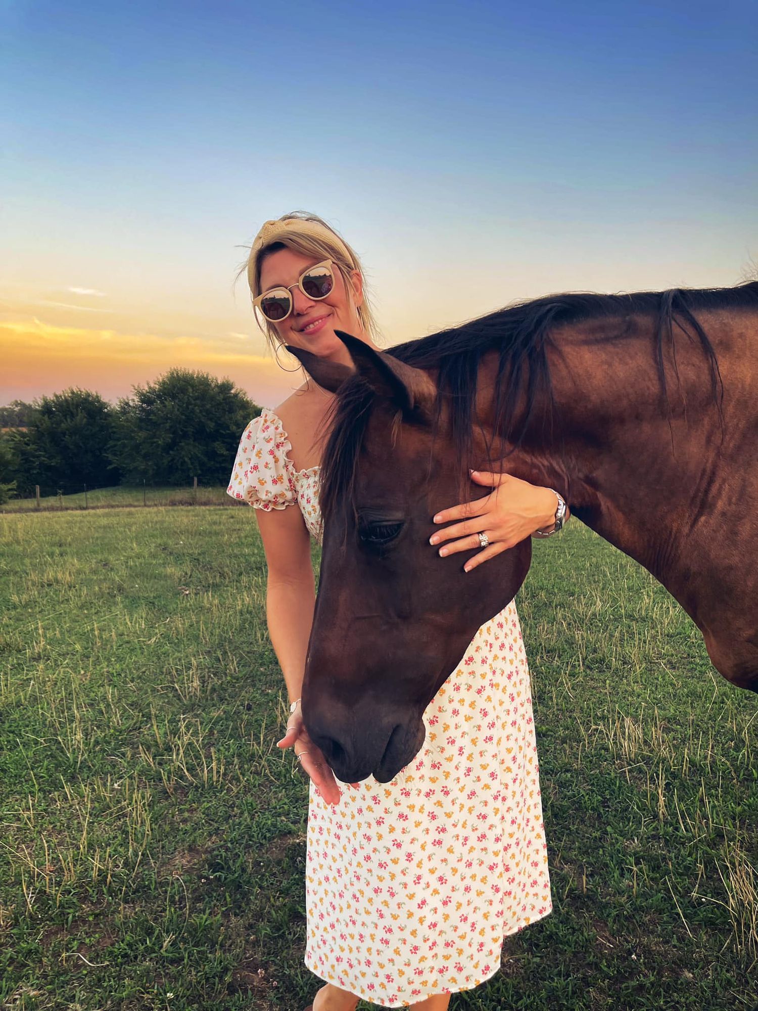 A person in a polka dot dress poses with a brown horse in a sunlit field at sunset.