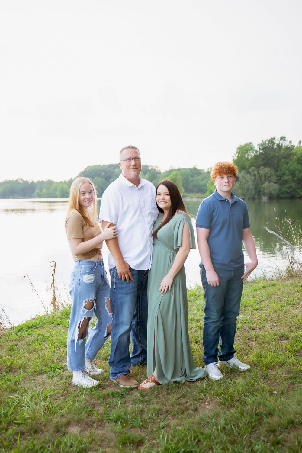Family of four poses together on grassy slope near lake during sunset.