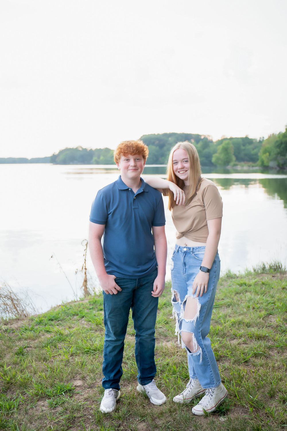 A casual outdoor portrait by a lake with two people standing on a grassy shore.