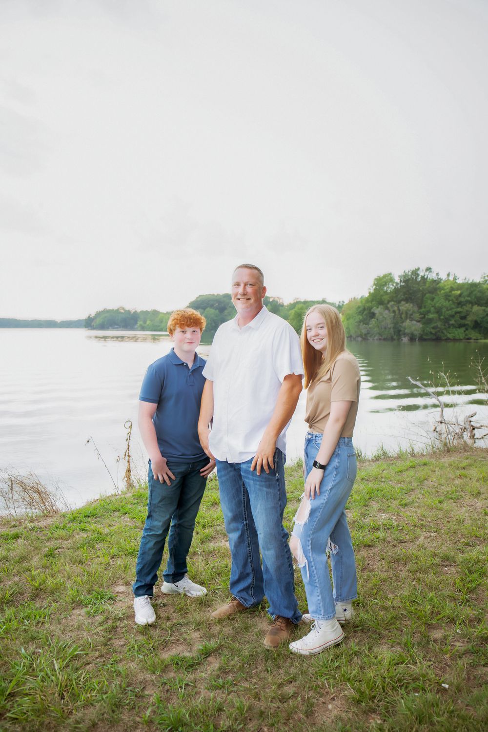 Three people stand together on a grassy lakeside bank during a family photoshoot.
