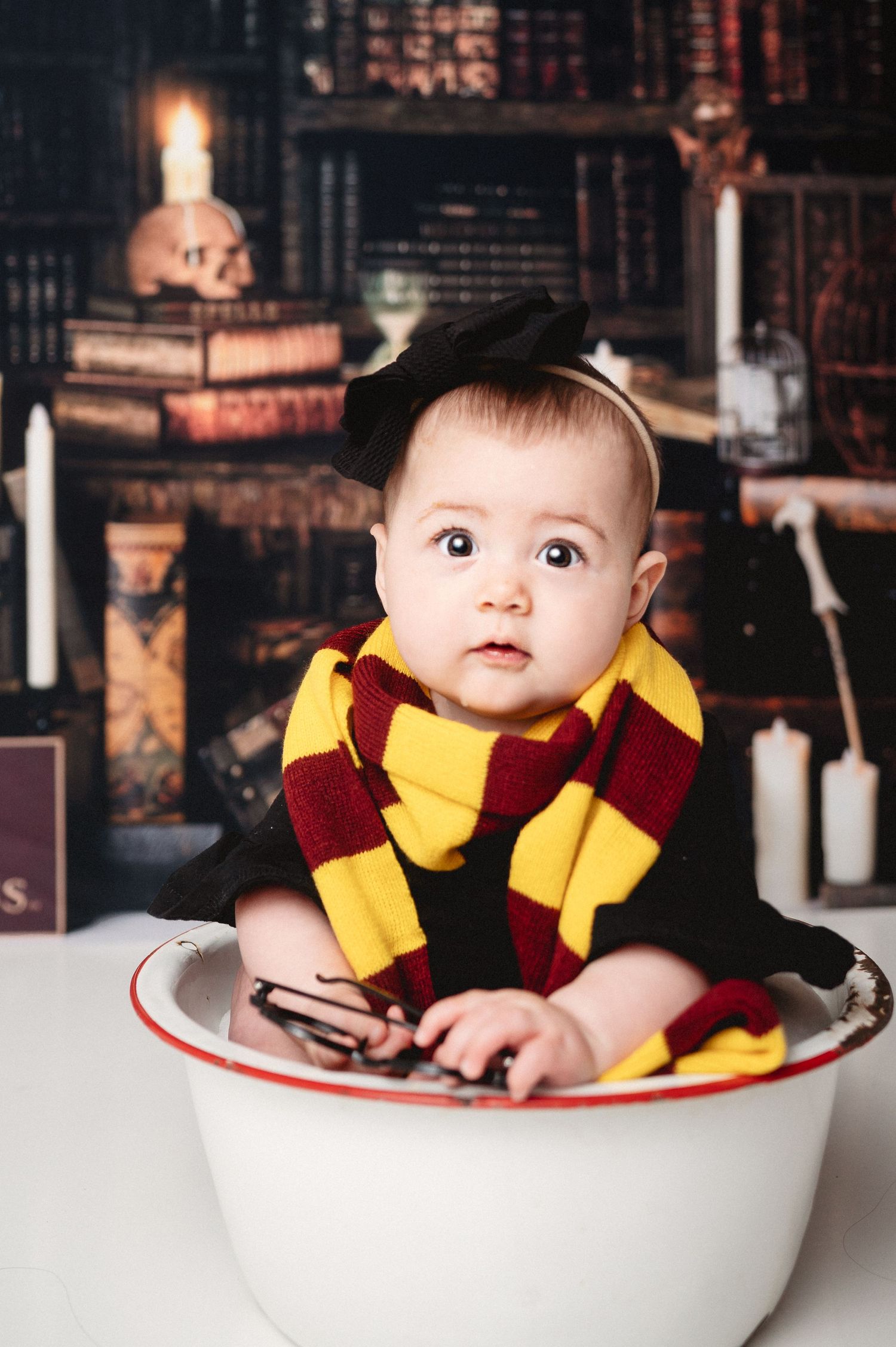 Baby in Gryffindor scarf sitting in white basin with magical library background.