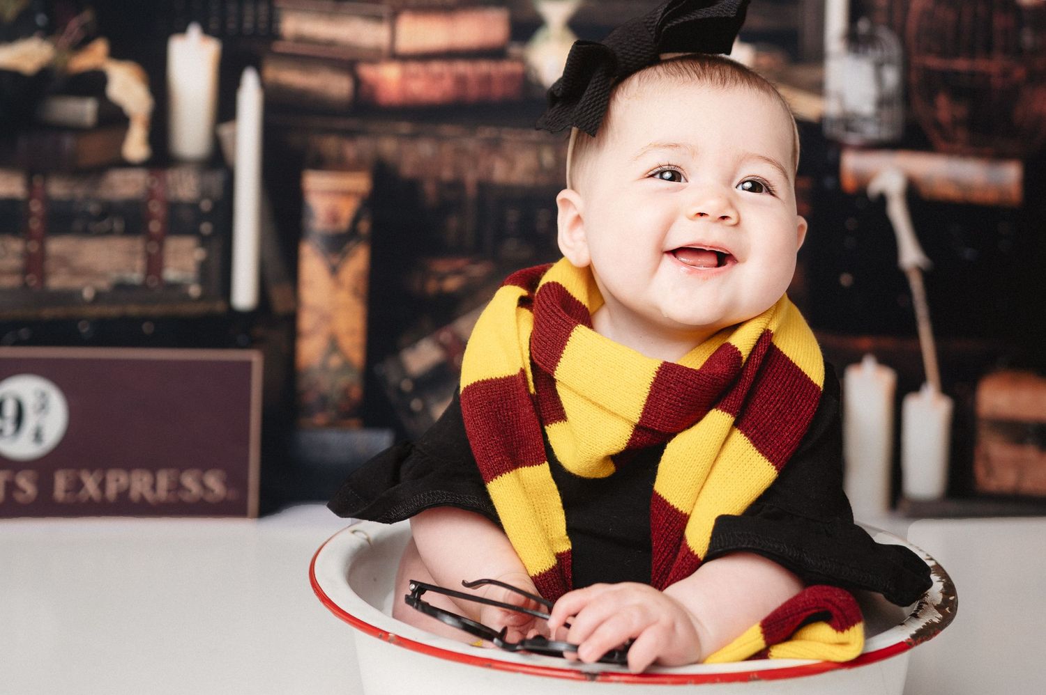 Baby wearing Gryffindor scarf smiles while sitting in white basin during photo session.