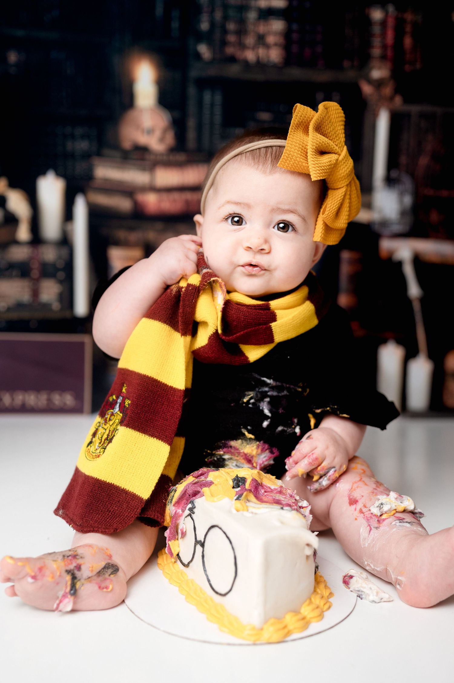 Baby in Gryffindor colors with yellow bow reaching for decorated Harry Potter cake.