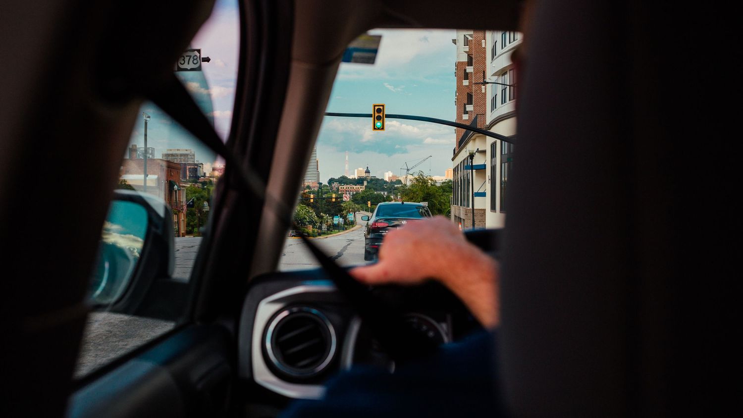 View from inside a car showing a driver at the wheel approaching a green traffic light in a city.