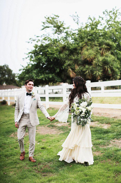 A romantic outdoor wedding moment with a couple on grassy field by white fence, bride holding elegant white floral bouquet.