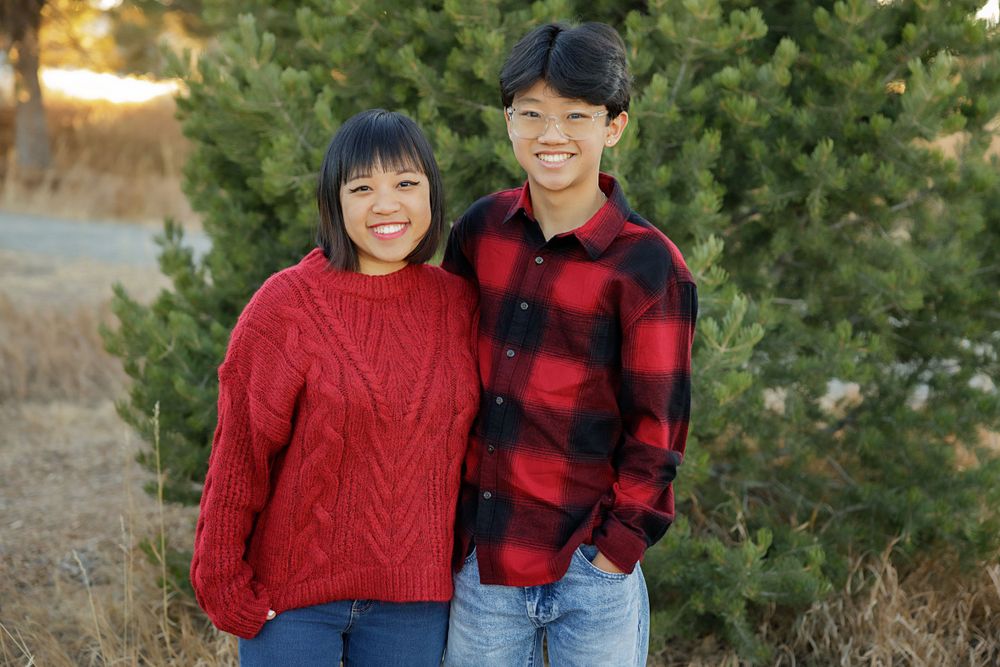 Two people in warm winter clothing smiling together in front of evergreen trees.