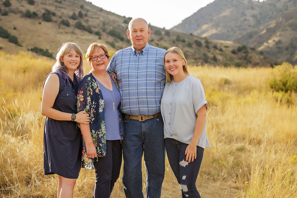 Family group portrait in casual clothing standing together in golden mountain field at sunset.