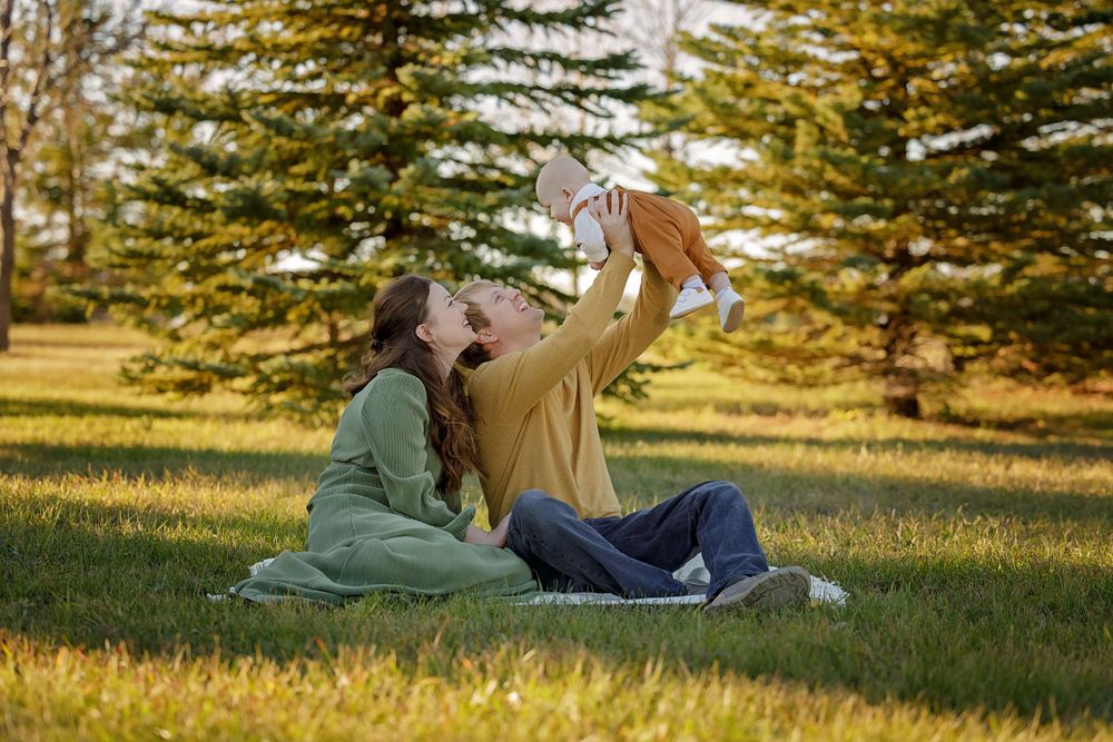 Couple lying on grass playing with baby under evergreen trees.