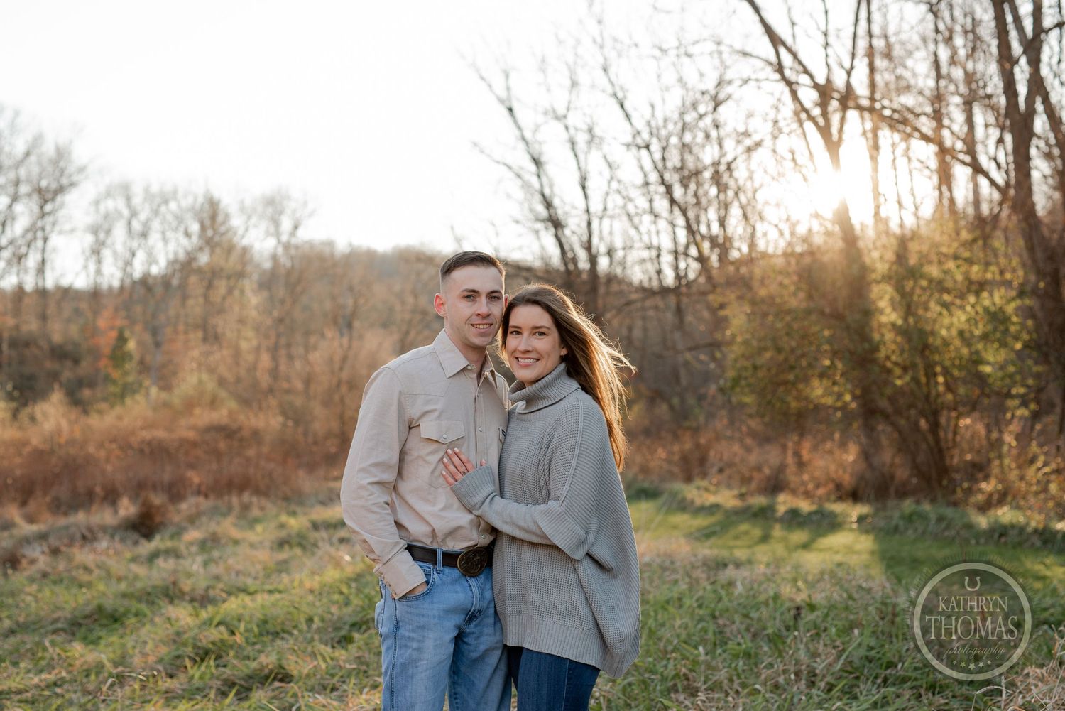 A couple embraces in a fall field during sunset wearing casual attire against a backdrop of bare trees.