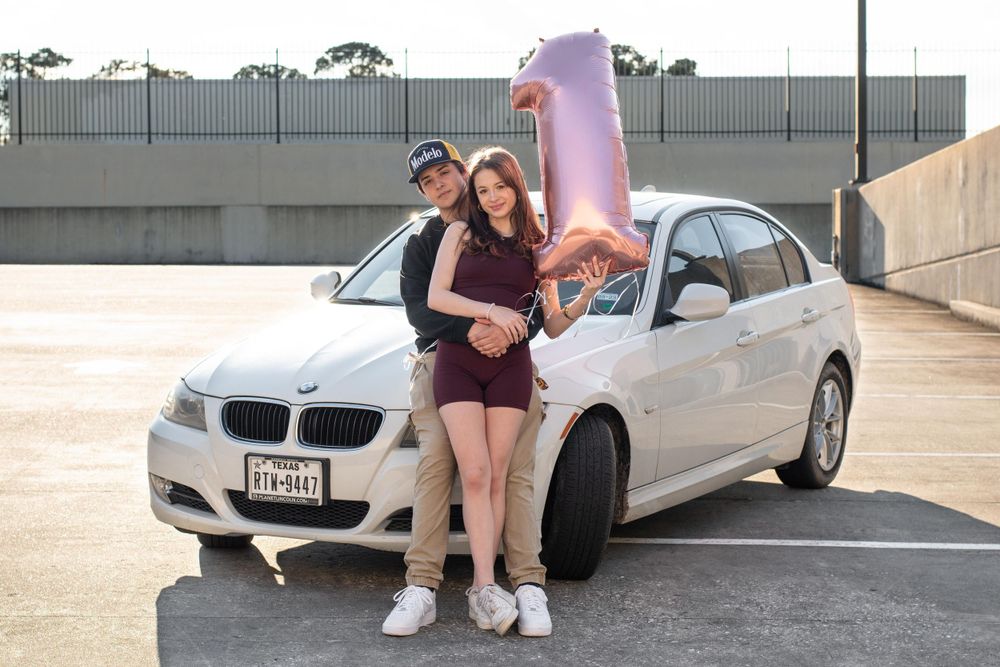 Couple posing with a white BMW and pink balloon in a parking garage.