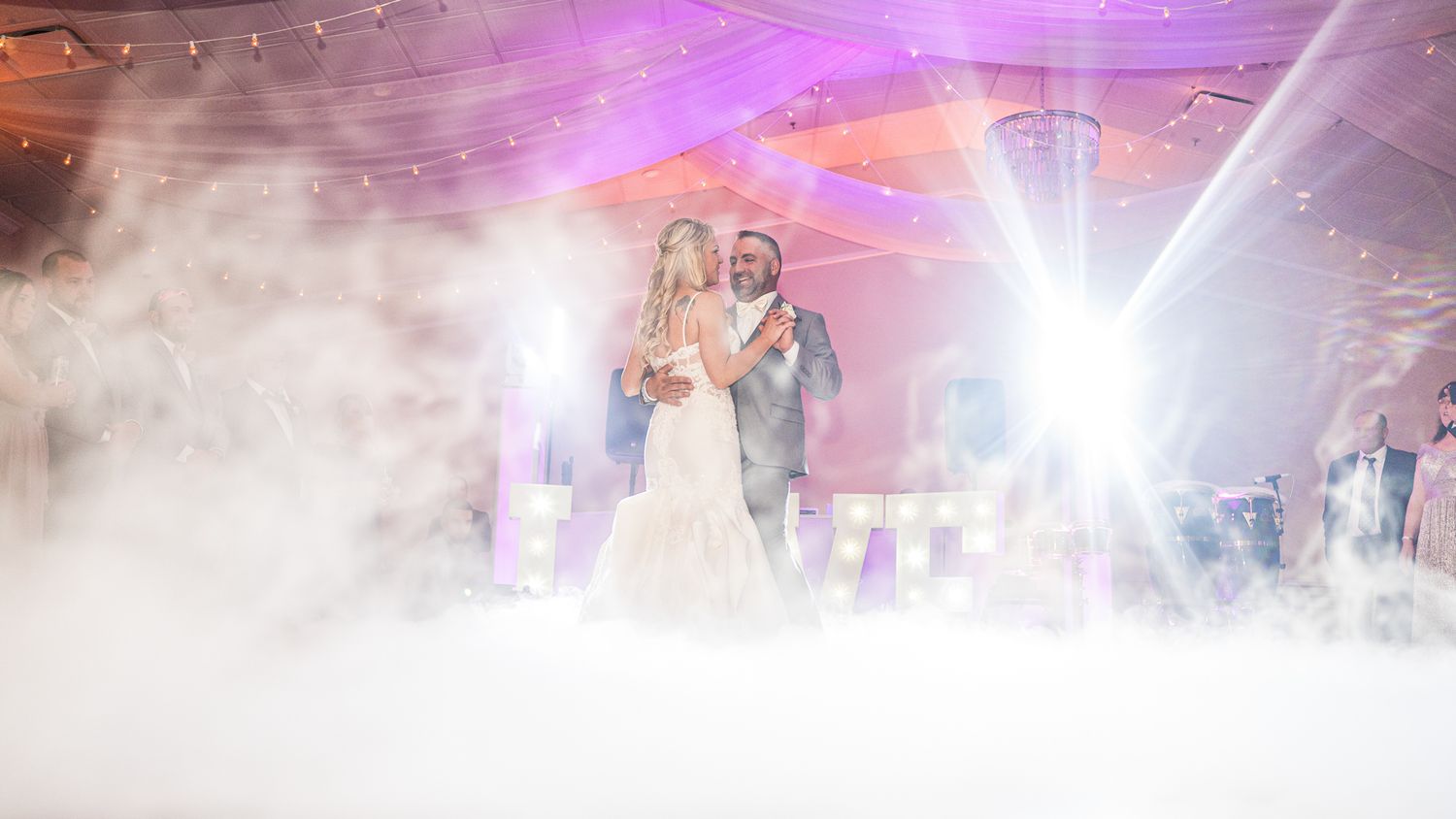 Soulful gaze as newlyweds share their first dance amidst colorful lights and swirling mist.