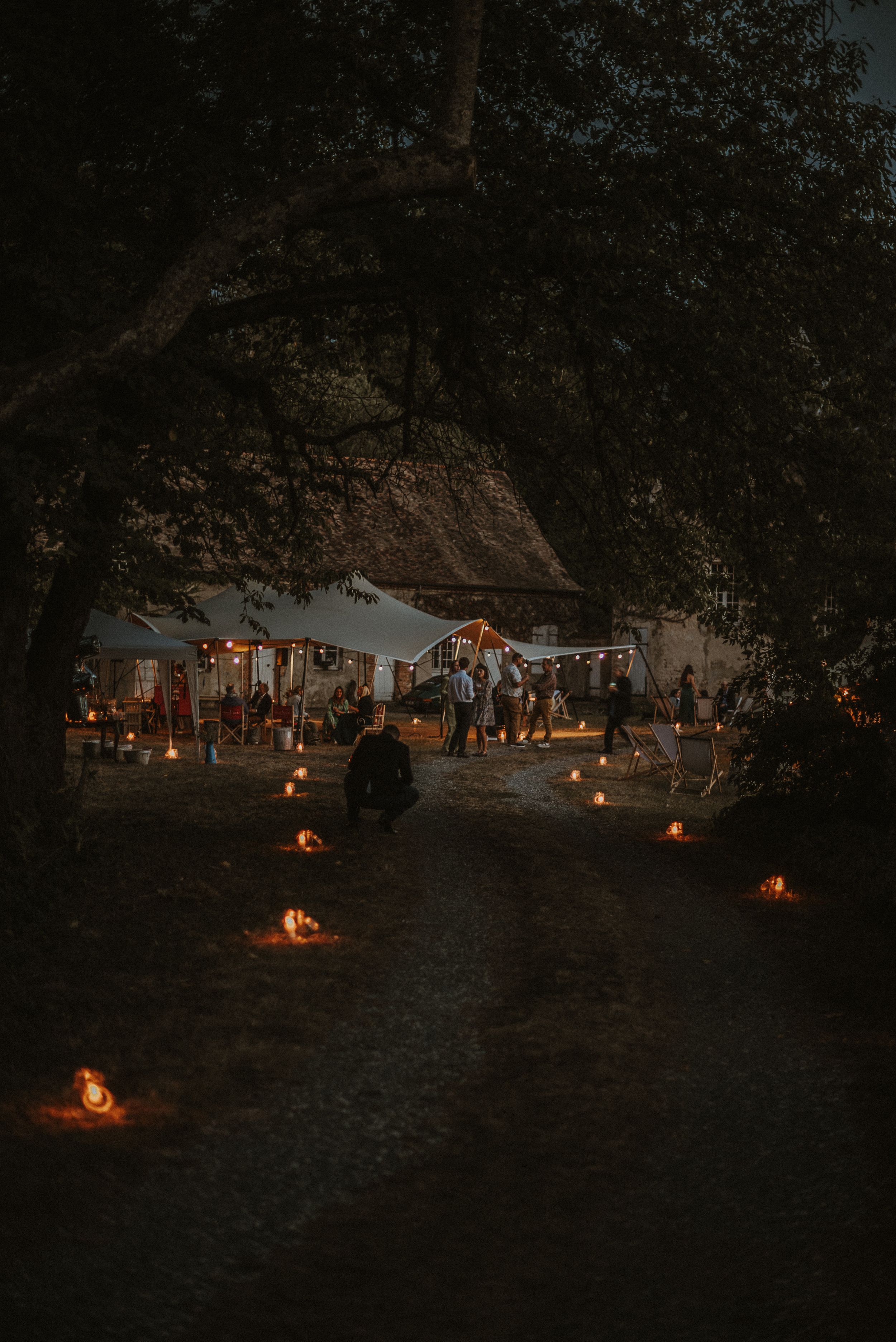 Candlelit pathway leading to an evening outdoor wedding reception under trees.