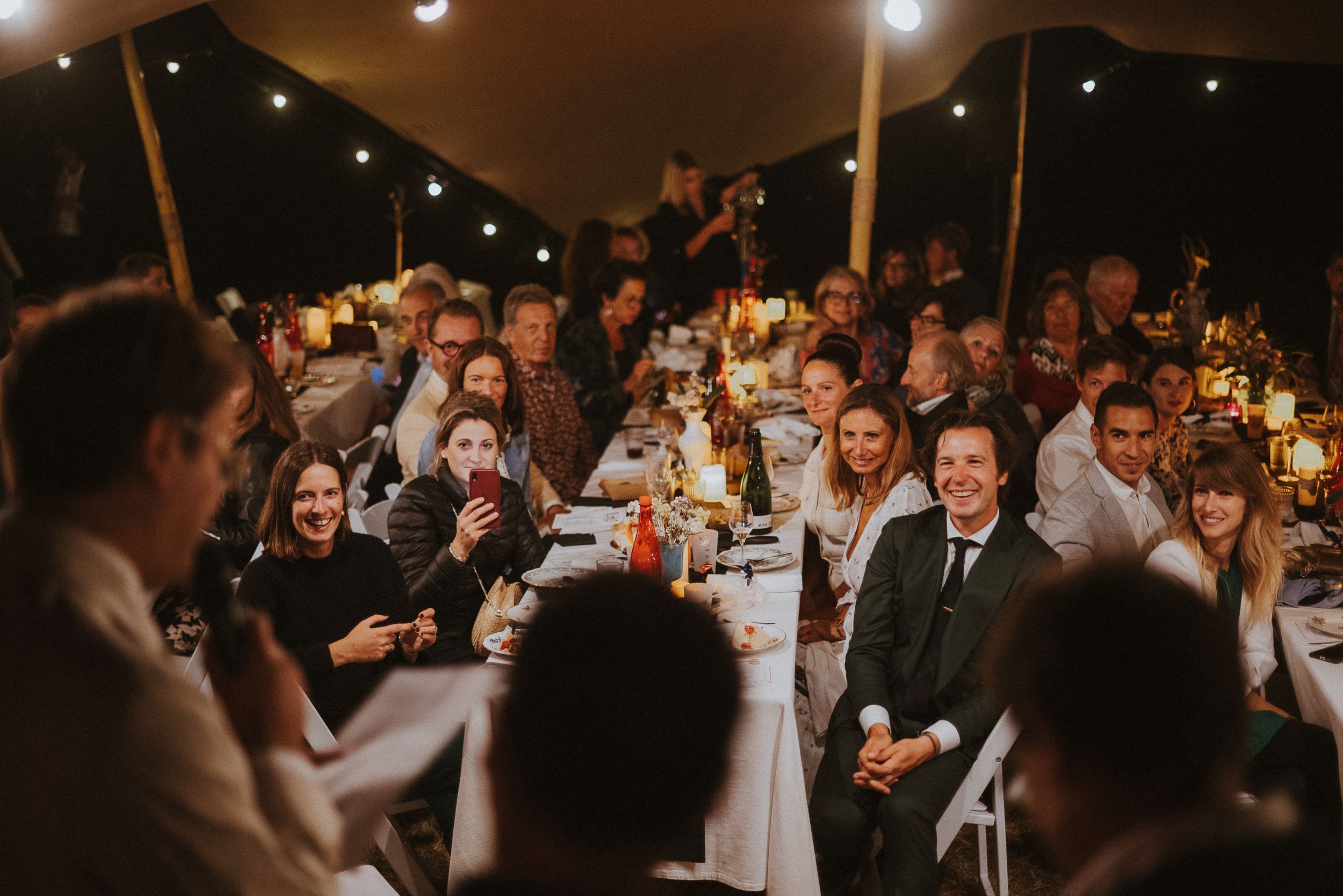 Wedding reception dinner guests seated at long tables under a tent with ambient lighting.