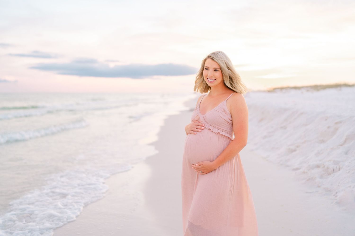 Expectant mother in pink dress standing on beach at sunset with waves in background.