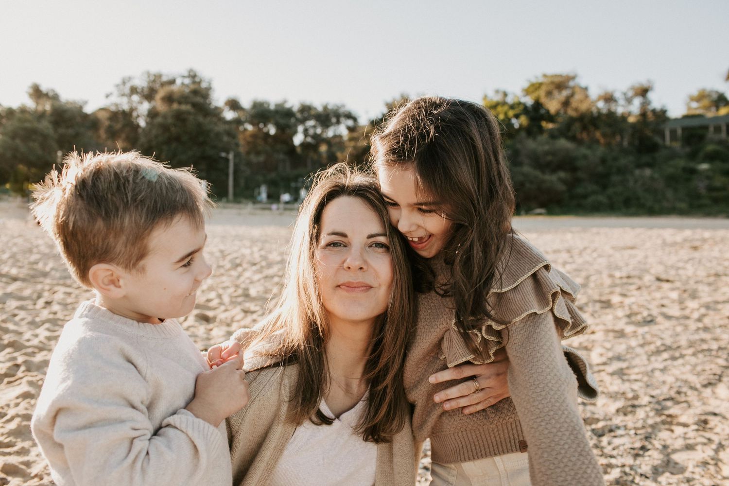 Smiling mother embraced by two young children on sandy beach in warm natural light.
