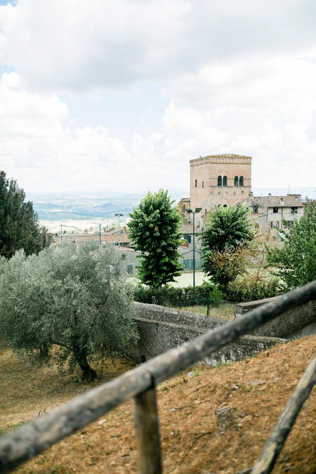 Historic stone tower and buildings perched on hilltop with olive trees in foreground.