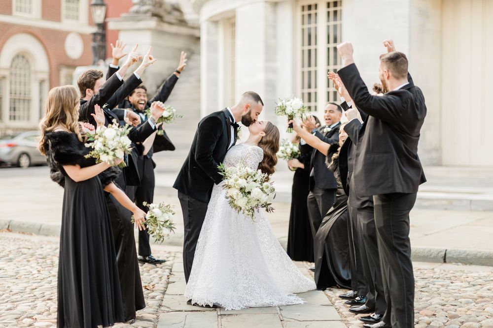 Wedding party in black attire celebrates with raised arms in a historic courtyard.