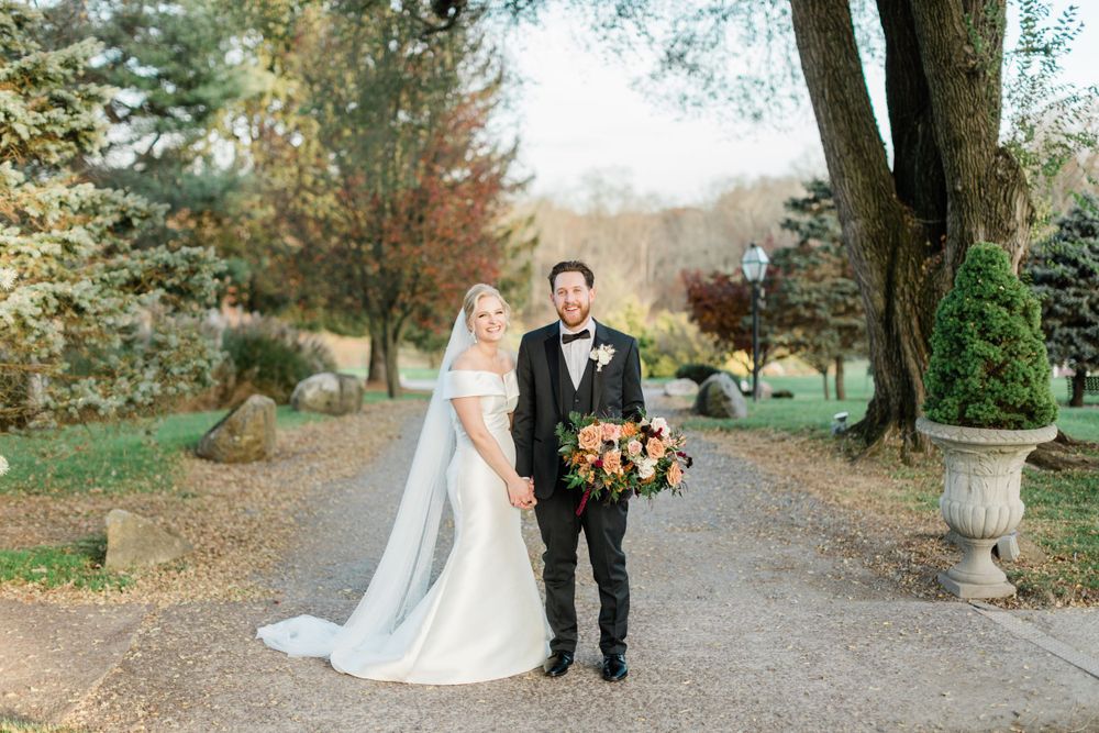 Fall wedding portrait on tree-lined path with couple holding autumn bouquet.