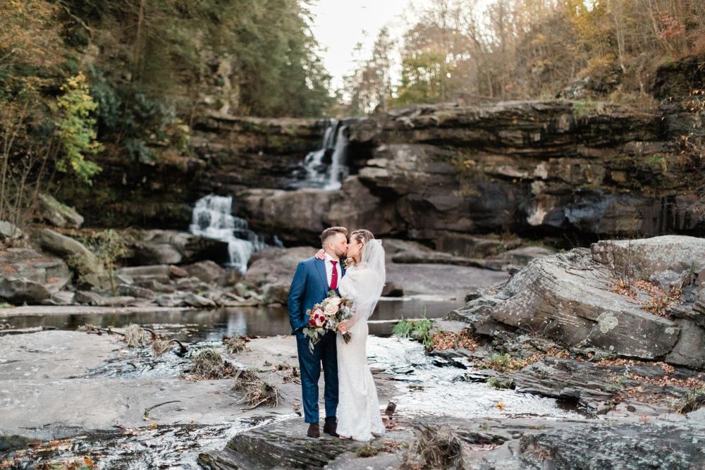 Couple poses in front of a cascading waterfall on rocky terrain during their wedding day.