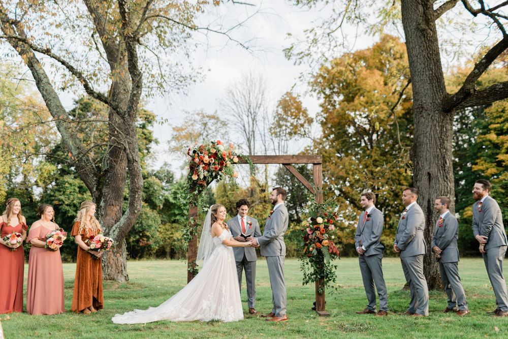 Outdoor wedding ceremony under a floral arch with coral-dressed bridesmaids and groomsmen.