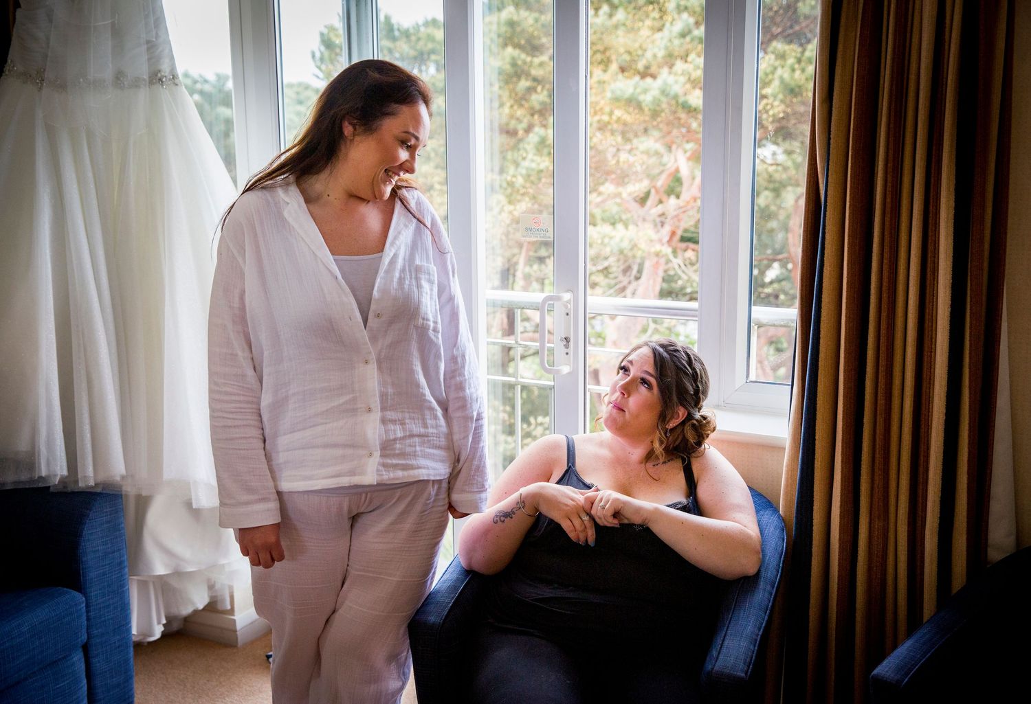 A relaxed pre-wedding moment in a bright room with wedding dress visible in background.