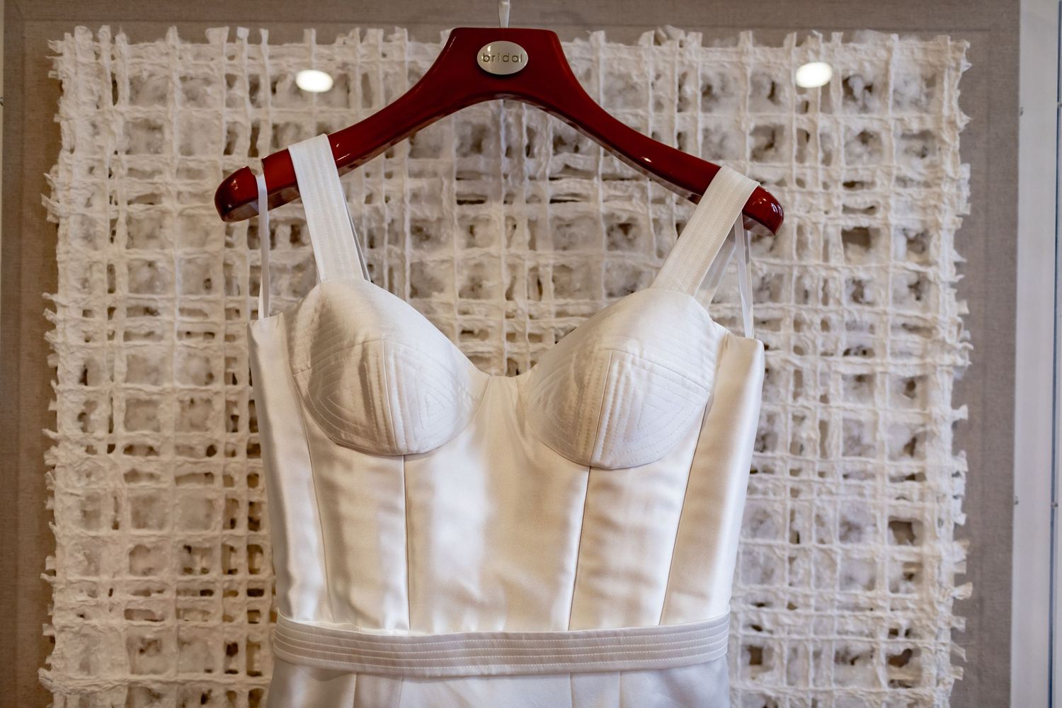 A close-up of a white wedding dress bodice on a red hanger against a textured background.