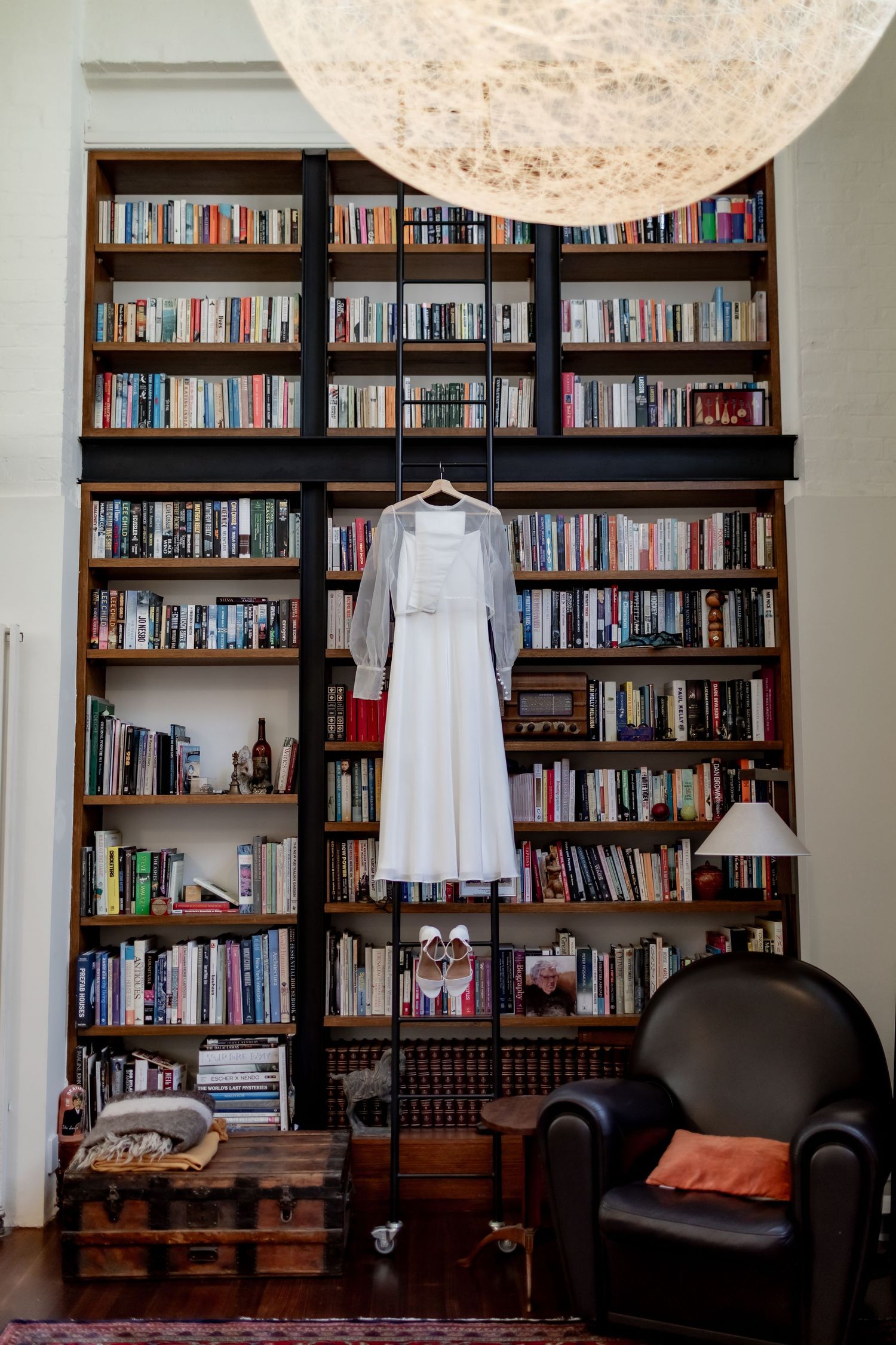 A white wedding dress hanging on a floor-to-ceiling bookshelf with a large pendant light.