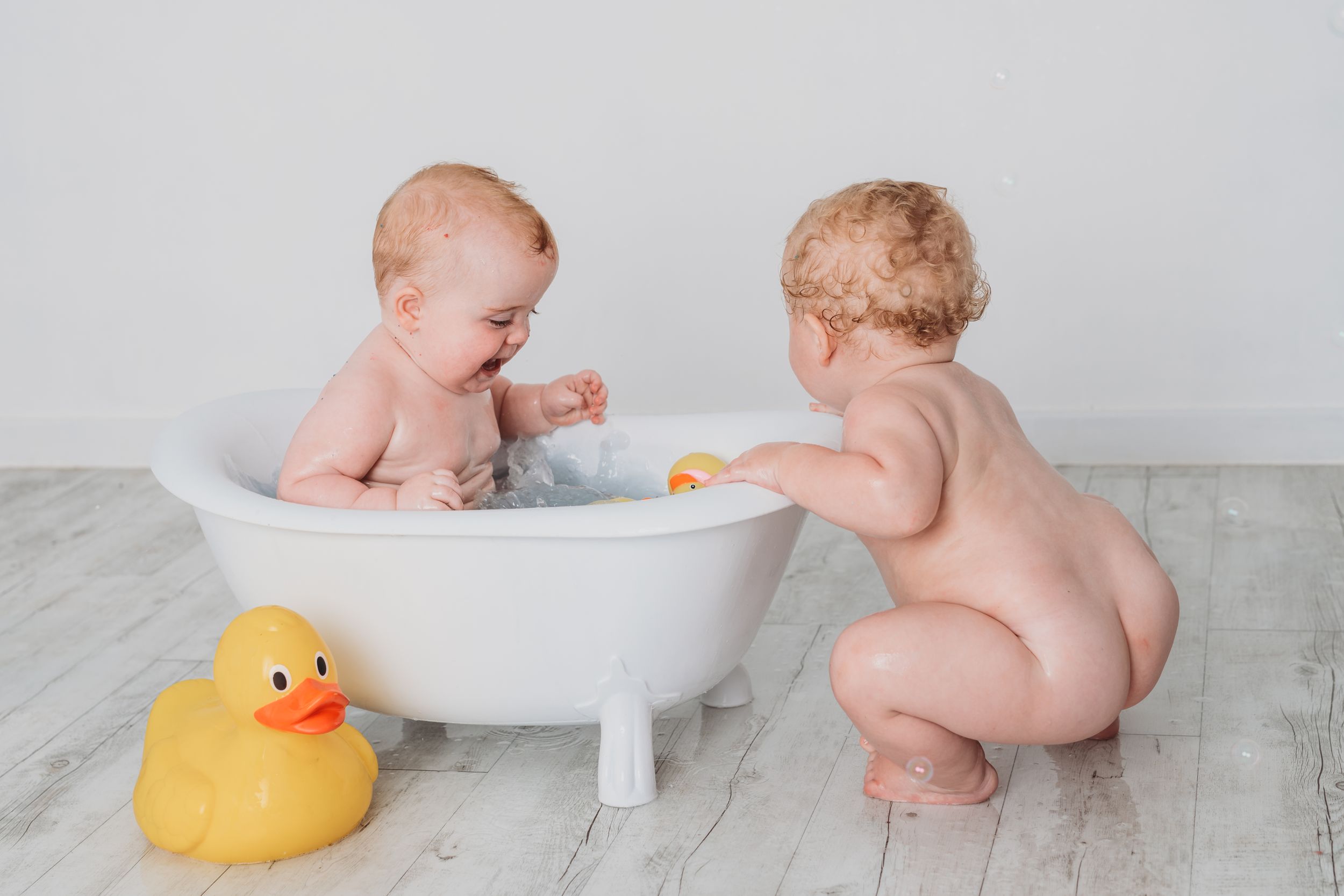 Two babies play with rubber duck near small white bathtub on wooden floor.