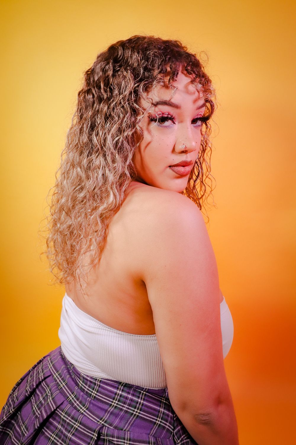 Young woman with curly blonde hair wearing white crop top and plaid skirt on yellow background.