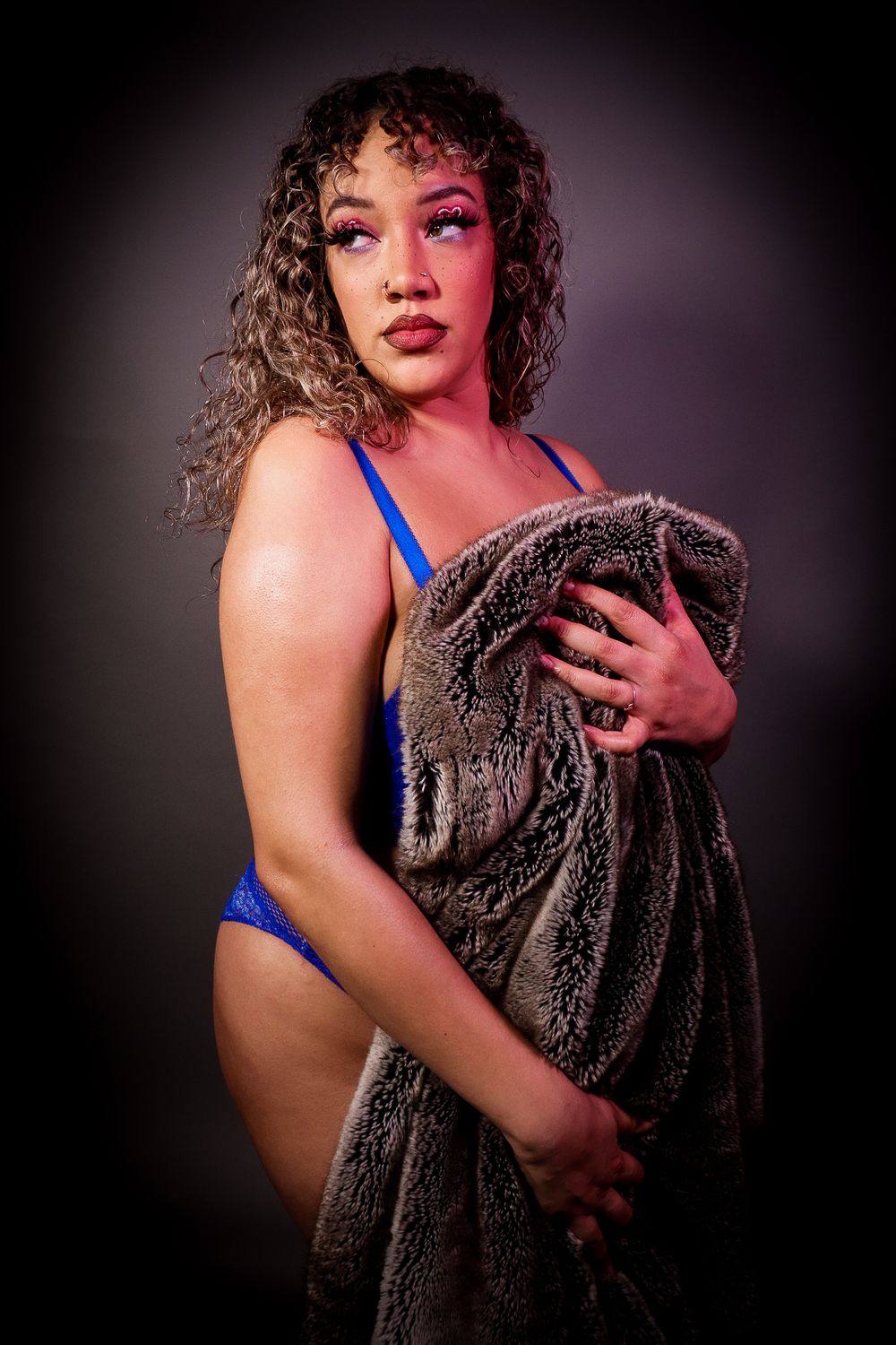 Curly haired woman in blue bikini holding gray faux fur coat in dramatic studio lighting.