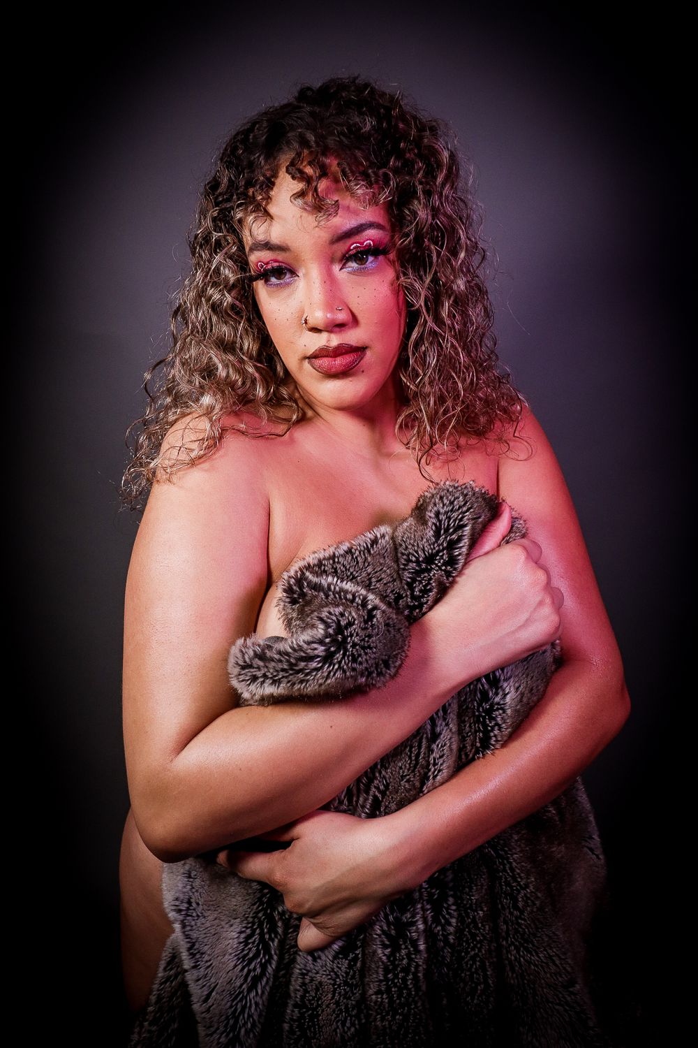 Curly haired woman with bold pink makeup holding gray faux fur coat in moody studio lighting.